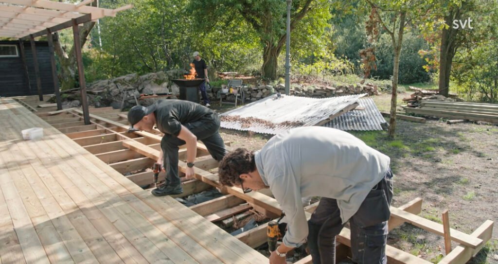 Stiligt - Två personer använder elverktyg för att bygga ett trädäck utomhus, medan en annan person står vid en eldstad i bakgrunden.
