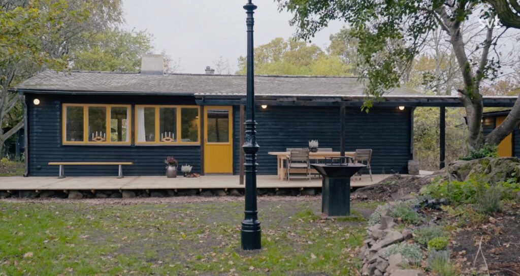 Stiligt - Ett svart trähus med gula dörrar och fönster, en veranda med uteservering och en lyktstolpe på gården omgiven av träd och gräs.