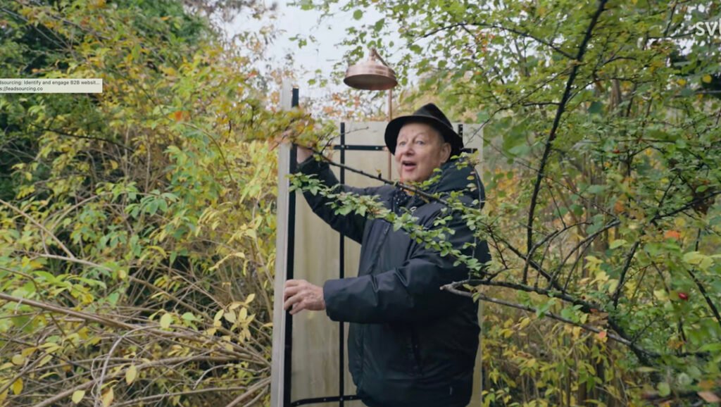 Stiligt - En man i utomhuskläder står i ett tätt lövverk och håller i en utomhusduscharmatur omgiven av lövgrenar.