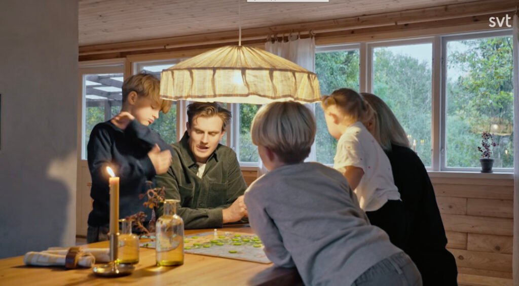 Stiligt - Fem personer, varav tre barn och två vuxna, sitter runt ett träbord och spelar ett brädspel i ett varmt upplyst rum med stora fönster och levande ljus.