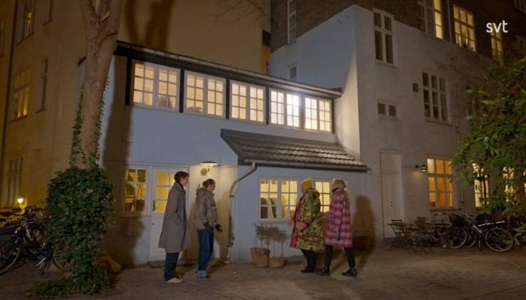 Stiligt - Fyra personer står och pratar utanför ett litet, ljusblått hus på kvällen, med lampor tända inomhus och cyklar parkerade i närheten.