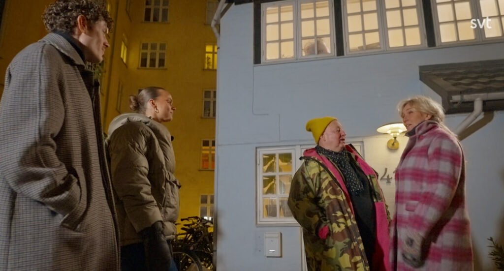 Stiligt - Fyra personer i vinterjackor står utanför en ljusblå byggnad på kvällen och pratar. Två står tätt intill varandra, medan två andra tittar på. Ett fönster ovanför är upplyst.