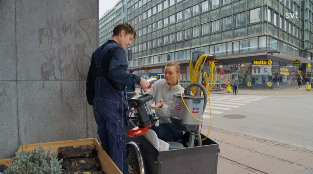 Stiligt - En man i overall räcker över ett föremål till en kvinna som sitter på en lastcykel på en trottoar i en stad, med butiker och fotgängare i bakgrunden.