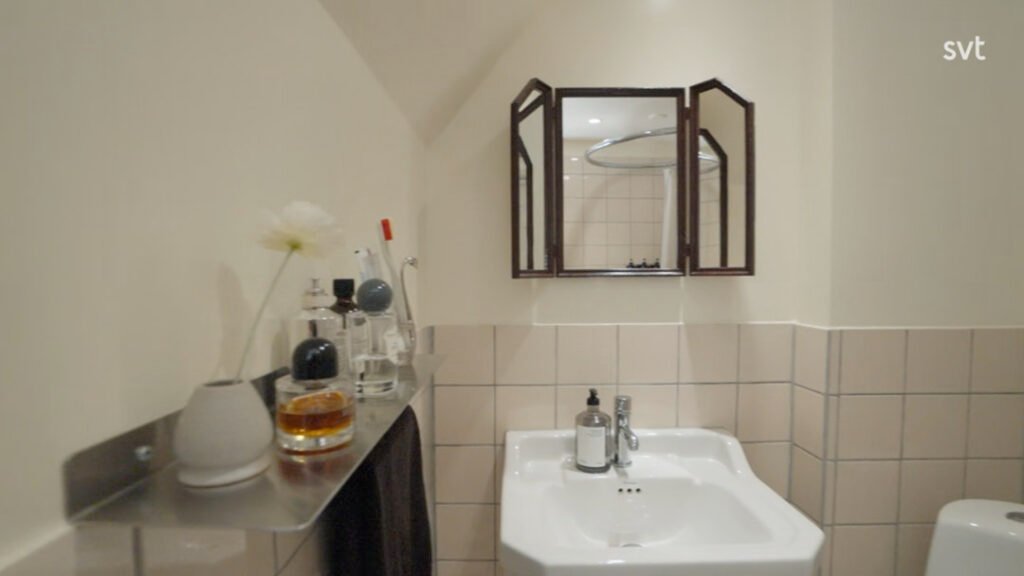 Stiligt - Ett badrum med handfat, tvålpump och spegelskåp; på en metallhylla finns flaskor, en vas med en blomma och en tandborste. Kaklade nedre väggar och gräddfärgade övre väggar är synliga.