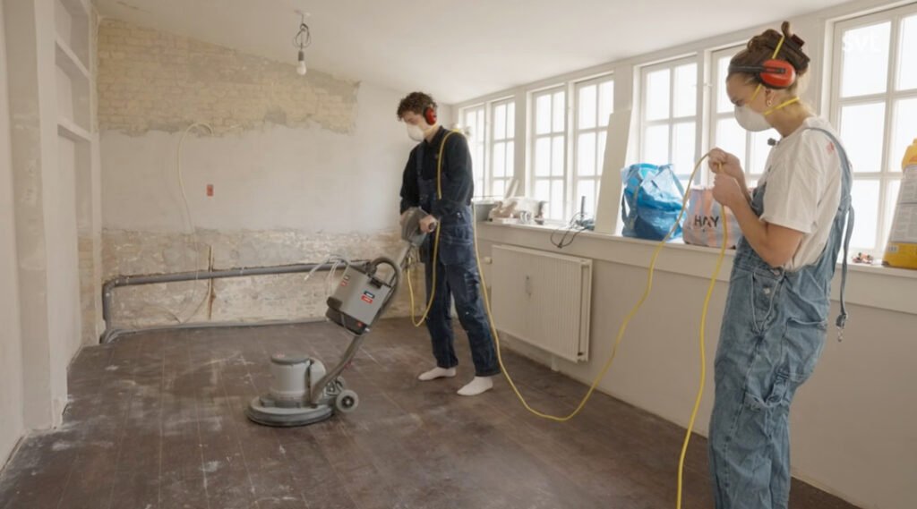 Stiligt - Två personer iförda skyddsmask och hörselskydd slipar ett trägolv i ett ljust, delvis renoverat rum.