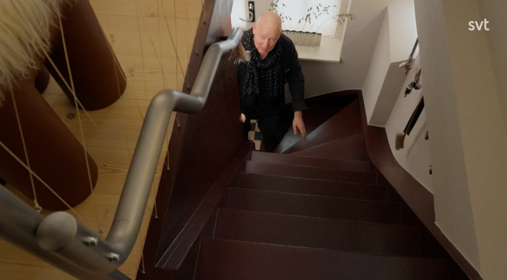 Stiligt - En äldre person med vitt hår går uppför en spiraltrappa i mörkt trä i en väl upplyst, modern interiör.