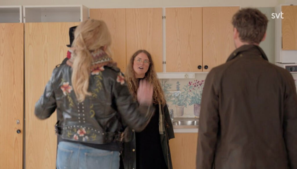 Stiligt - Tre personer står och pratar i ett kök med träskåp; en kvinna är vänd mot kameran medan de andra två har ryggen mot.