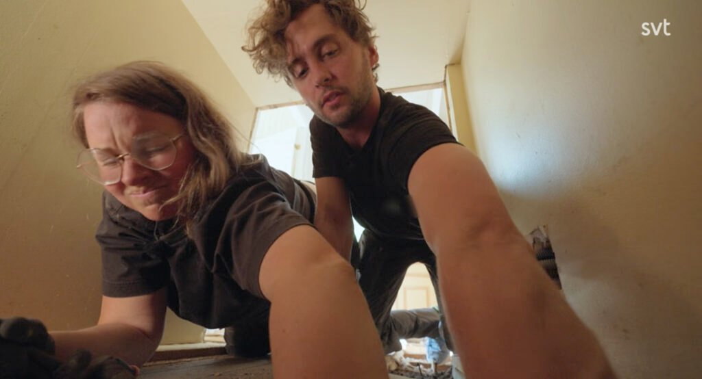 Stiligt - Två personer i fritidskläder arbetar tätt tillsammans på golvet nära en vägg och verkar reparera eller installera något i ett trångt inomhusutrymme.