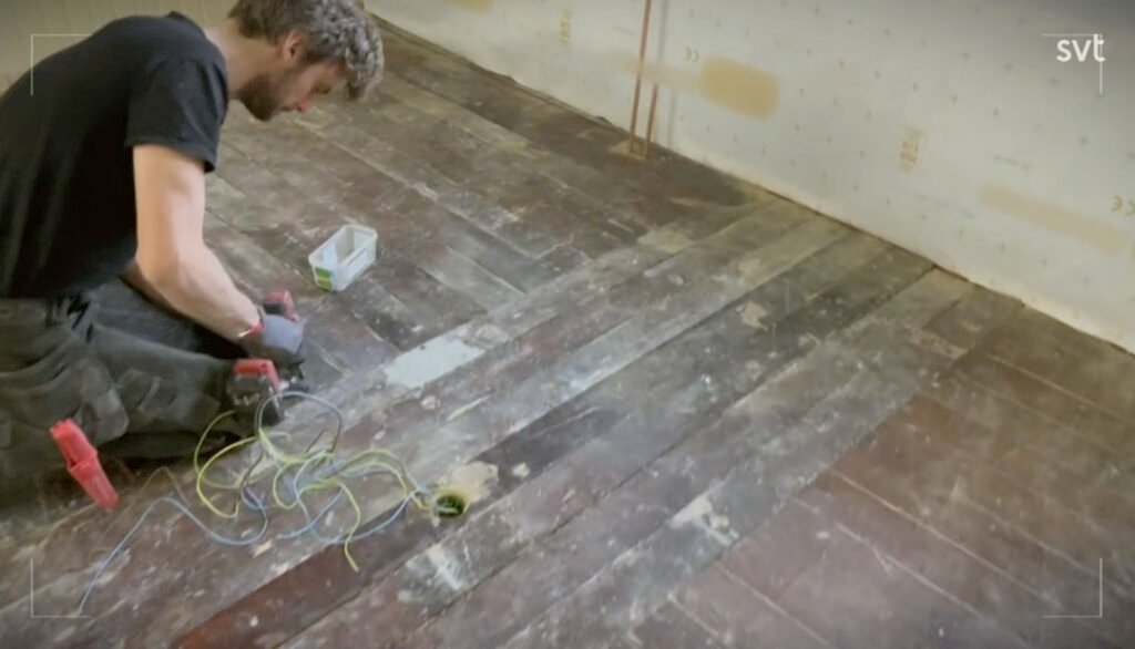 Stiligt - En person står på knä på ett slitet trägolv och arbetar med sladdar och verktyg i närheten av ett litet hål i golvet i ett ofärdigt rum.