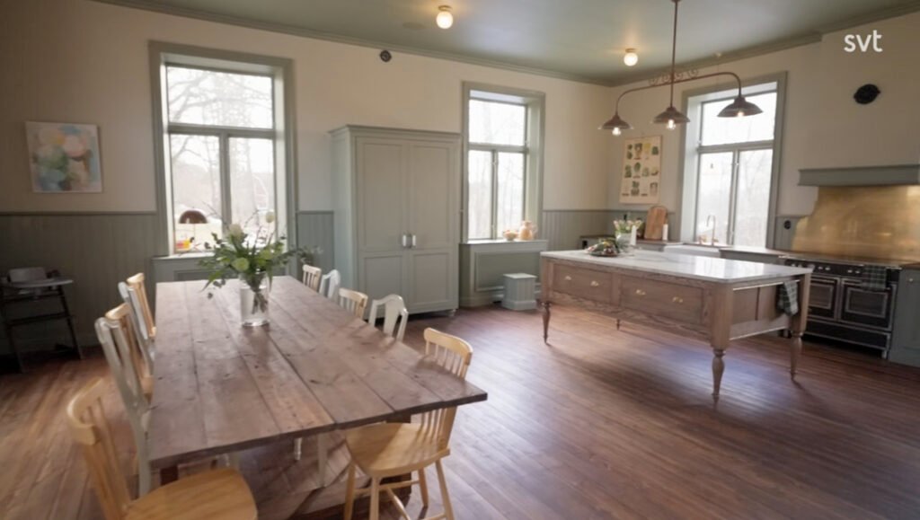 Stiligt - Ett rymligt kök med ett långt matbord i trä, ljusa stolar, stora fönster, en köksö och vitvaror i rostfritt stål.