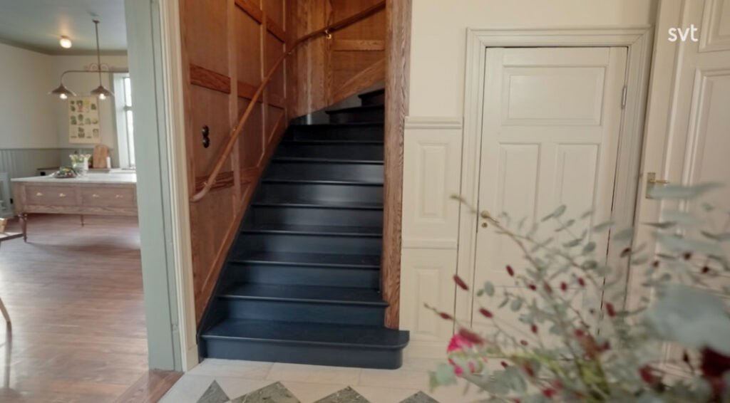 Stiligt - En svart trappa med ledstång i trä leder upp i en hall med cremefärgade dörrar och träpanel på väggarna; till vänster syns en matsal.