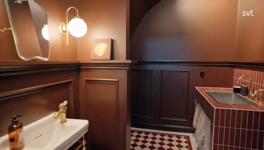 Stiligt - Ett badrum med bruna väggar, guldfärgade armaturer, ett vitt handfat, en kaklad bänk med svart handfat, en rund vägglampa och röd- och vitrutigt klinkergolv.