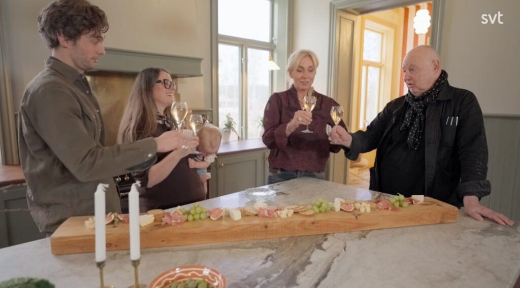 Stiligt - Fyra vuxna står runt en köksö med en lång charkuteribräda och håller vinglas i en skål; en kvinna håller en bebis.