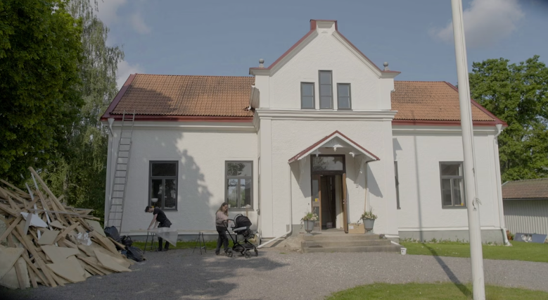 Stiligt - Ett vitt tvåvåningshus med rött tak, byggavfall till vänster och två vuxna med barnvagn som står nära entrén en solig dag.