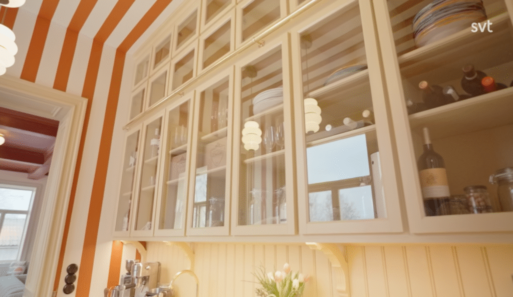 Stiligt - I köksskåpen med glasfront förvaras porslin, glas och flaskor. Väggarna har vertikala ränder i orange och vitt. Pendellampor och ett fönster är synliga.