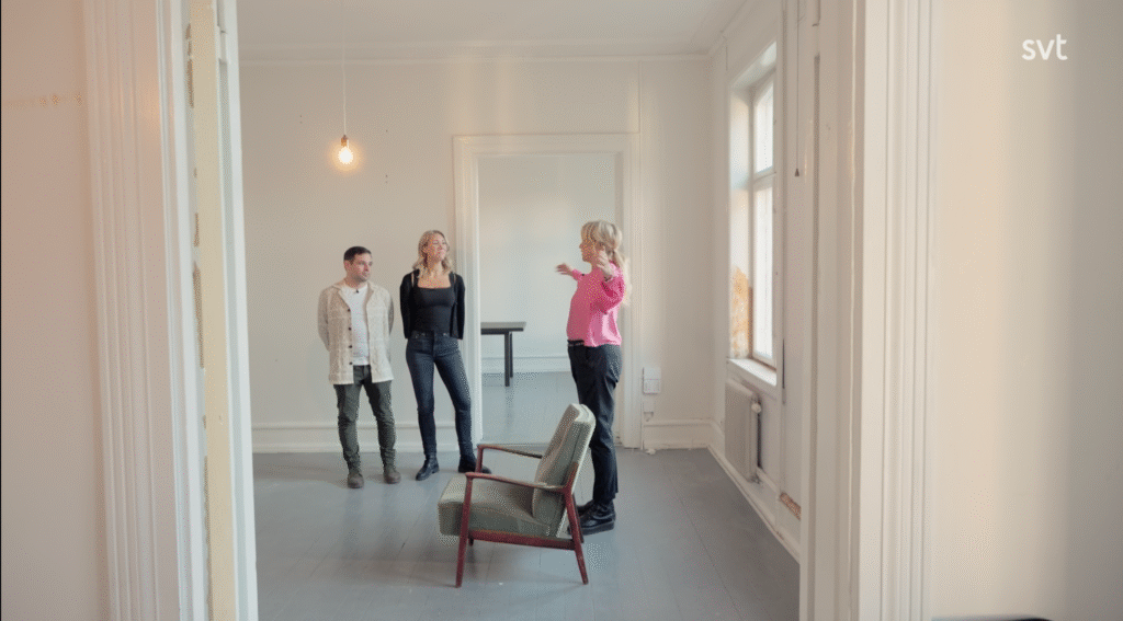 Stiligt - Tre personer står och pratar i ett i stort sett tomt rum med vita väggar och två stolar; en person gestikulerar medan de andra lyssnar.