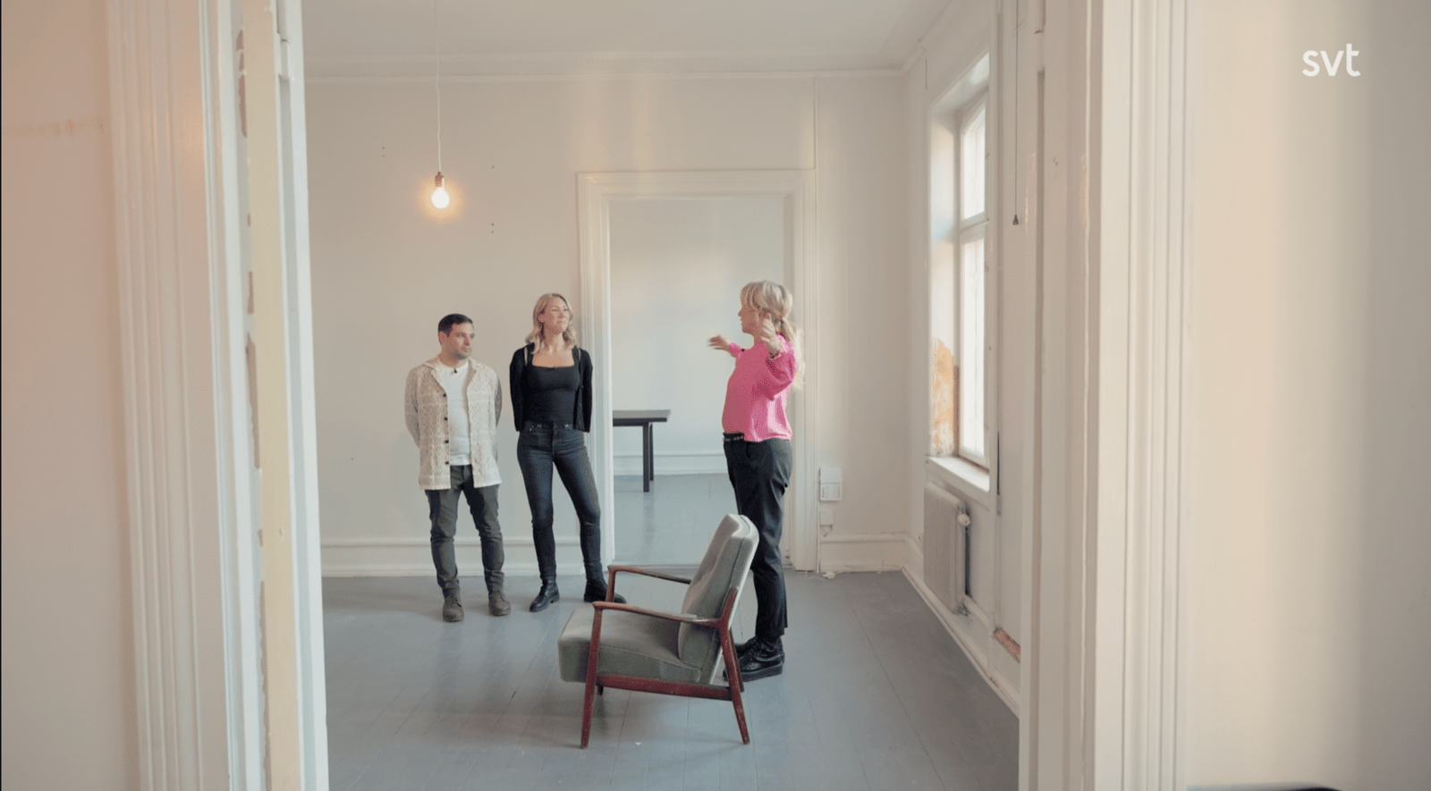 Stiligt - Tre personer står och pratar i ett i stort sett tomt rum med vita väggar och två stolar; en person gestikulerar medan de andra lyssnar.