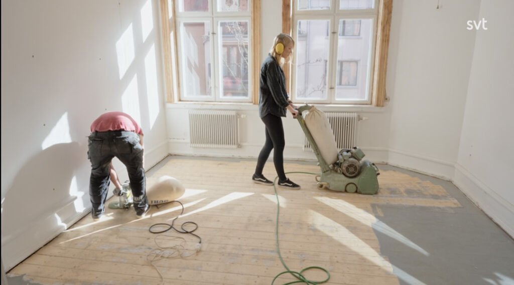 Stiligt - Två personer slipar ett trägolv i ett ljust, tomt rum; den ena använder en golvslipmaskin medan den andra arbetar nära väggen.