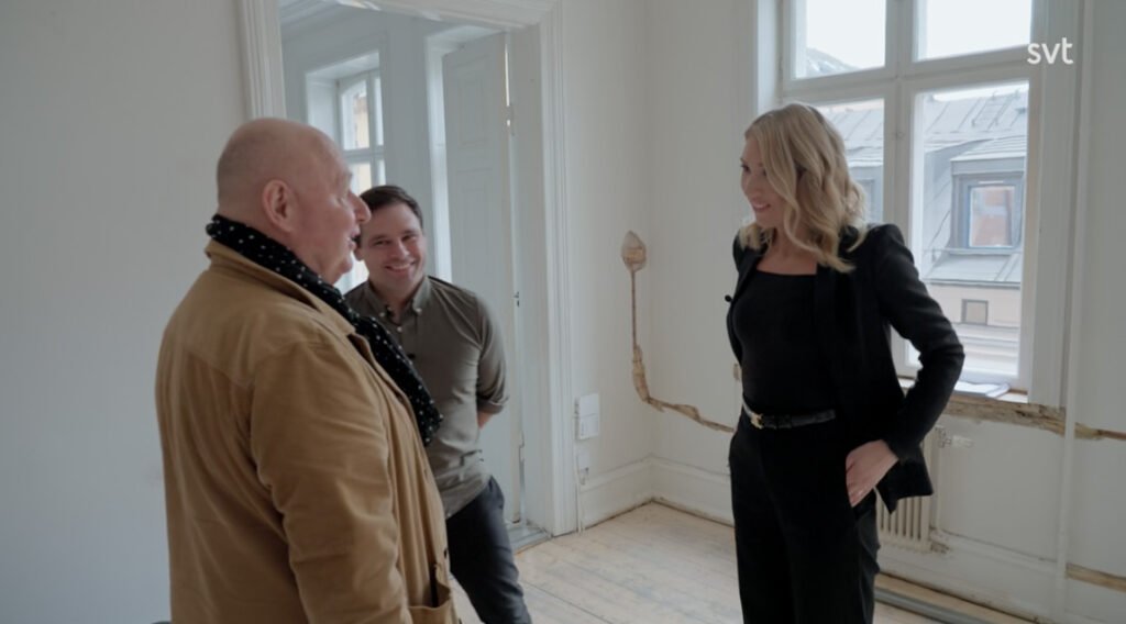 Stiligt - Tre personer står och pratar i ett ljust, omöblerat rum med stora fönster och synliga skador på väggarna.