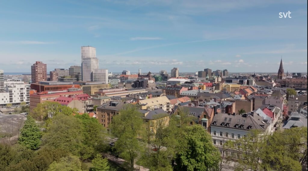 Stiligt - Flygfoto över en stadsbild med moderna och historiska byggnader, gröna träd i förgrunden och en blå himmel ovanför.