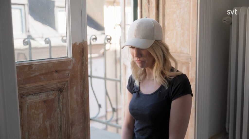 Stiligt - En kvinna med vit keps och svart skjorta står inomhus vid ett öppet fönster och tittar ner med ett neutralt ansiktsuttryck.