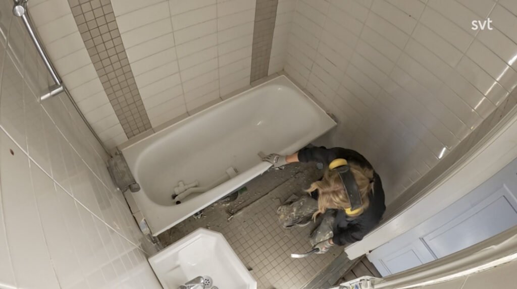 Stiligt - En person i skyddsutrustning tar bort kakel och skräp från ett badrumsgolv bredvid ett badkar, sett uppifrån.