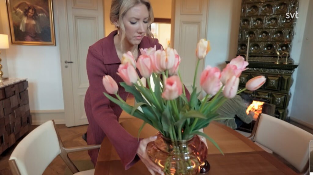 Stiligt - En kvinna i rödbrun klädsel arrangerar en vas med rosa tulpaner på ett matbord i trä i ett varmt upplyst rum.