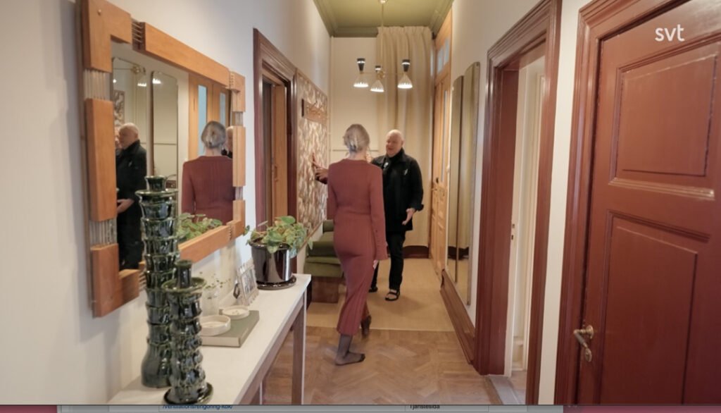 Stiligt - En kvinna i röd klänning går genom en korridor mot en äldre man som står nära en dörröppning; korridoren är dekorerad med speglar, en växt och en grön keramikskulptur.