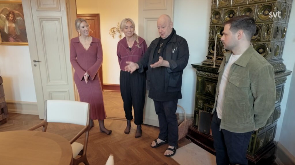 Stiligt - Fyra personer står och pratar i ett rum med trägolv och en dekorativ kakelugn. En person gestikulerar med sina händer medan de andra lyssnar.