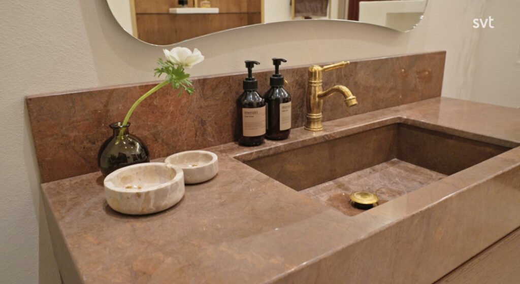 Stiligt - Ett badrumshandfat i brun sten med en guldkran, två tvålbehållare, en liten vas med en vit blomma och två stenfat på bänkskivan.