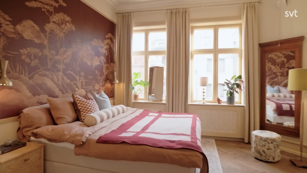 Stiligt - Ett sovrum med en stor säng, mönstrade kuddar, en rödbrun och beige filt, en tapet med ett naturmotiv, två fönster med gardiner, en golvspegel och krukväxter.