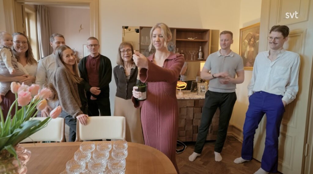 Stiligt - En kvinna öppnar en flaska i ett vardagsrum medan åtta personer, inklusive ett barn, tittar på. Champagneglas står uppställda på ett bord i förgrunden.