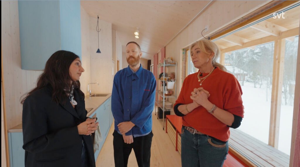 Stiligt - Tre personer står och samtalar i ett modernt, ljust kök med träväggar och stora fönster som visar ett snöigt landskap utanför.