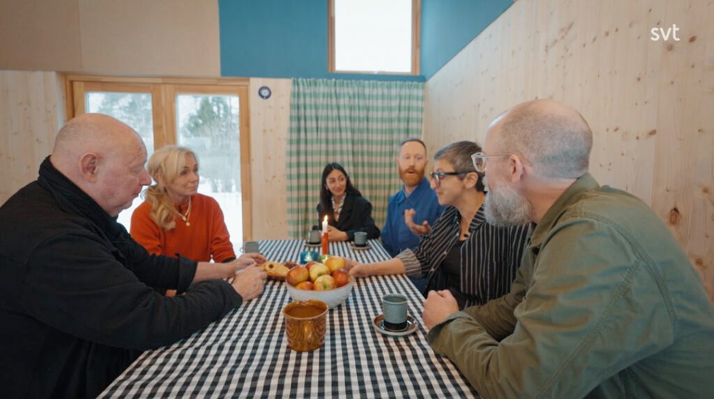 Stiligt - Sex vuxna sitter runt ett rutigt bord med frukt och koppar och samtalar i ett ljust rum med träpanel och stora fönster.