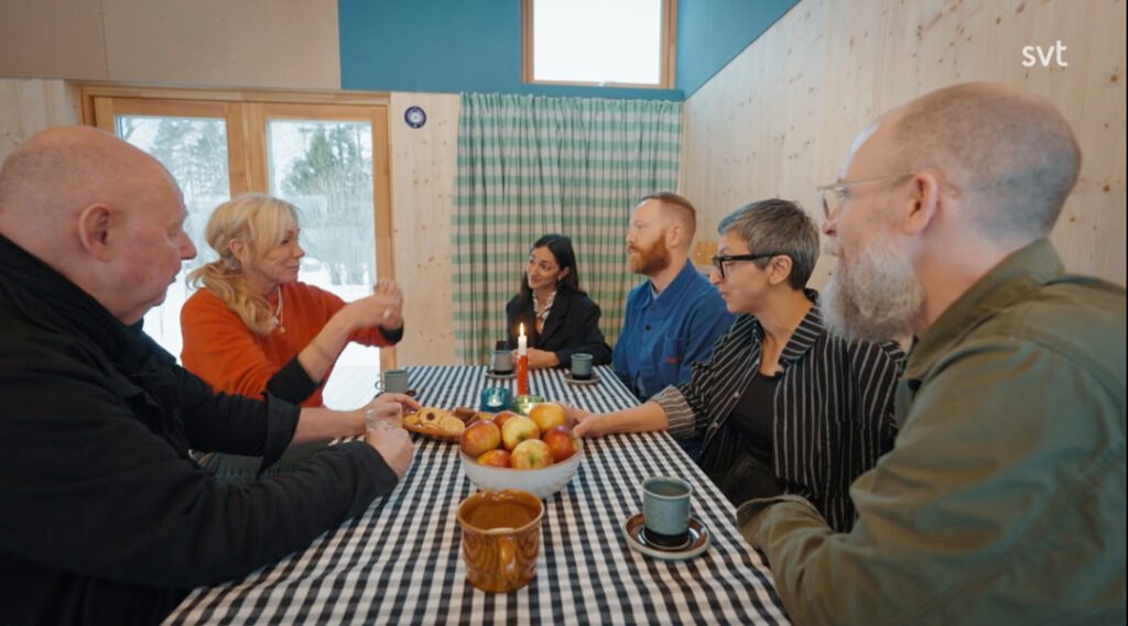 Stiligt - Sex vuxna sitter runt ett rutigt bord med frukt och kaffekoppar och samtalar i ett varmt upplyst rum med träväggar och fönster.