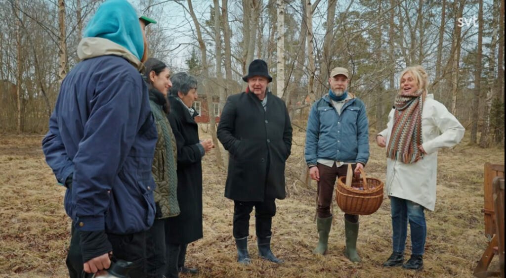 Stiligt - Sex vuxna står utomhus på en gräsbevuxen, lövtäckt yta i kalla kläder, med kala träd i bakgrunden; en person håller i en korg.