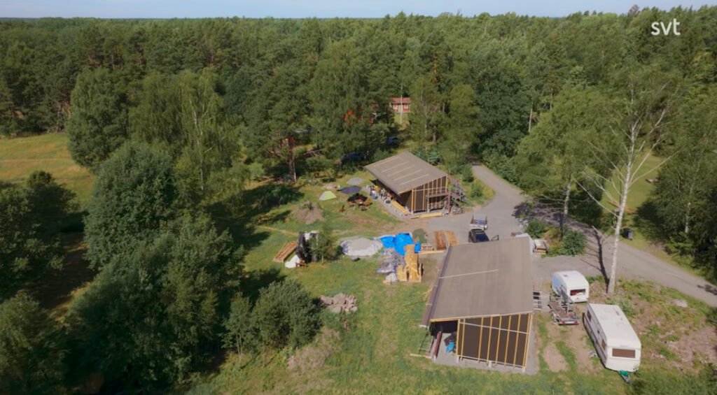 Stiligt - Flygfoto över två moderna träbyggnader omgivna av träd, med en parkerad vit skåpbil, släpvagn och spridda byggmaterial i närheten.