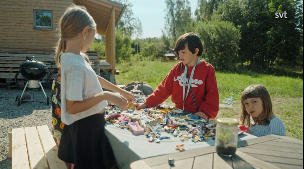 Stiligt - Tre barn leker med legobitar på ett bord utomhus i närheten av en trästuga en solig dag.