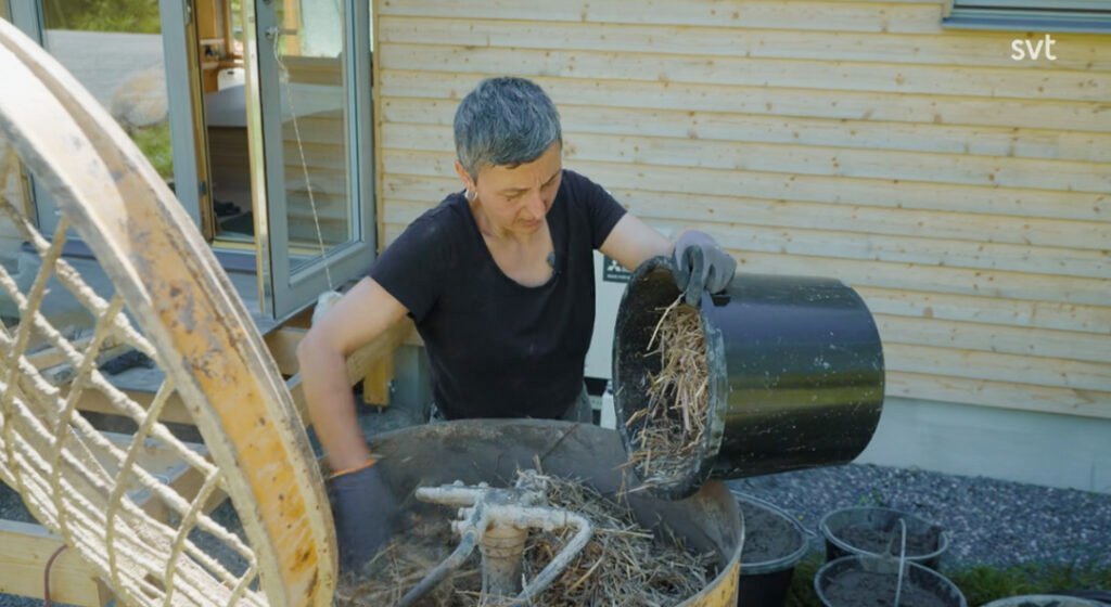 Stiligt - En person iförd handskar häller halm och organiskt material från en svart hink i en cementblandare utanför en träbyggnad.