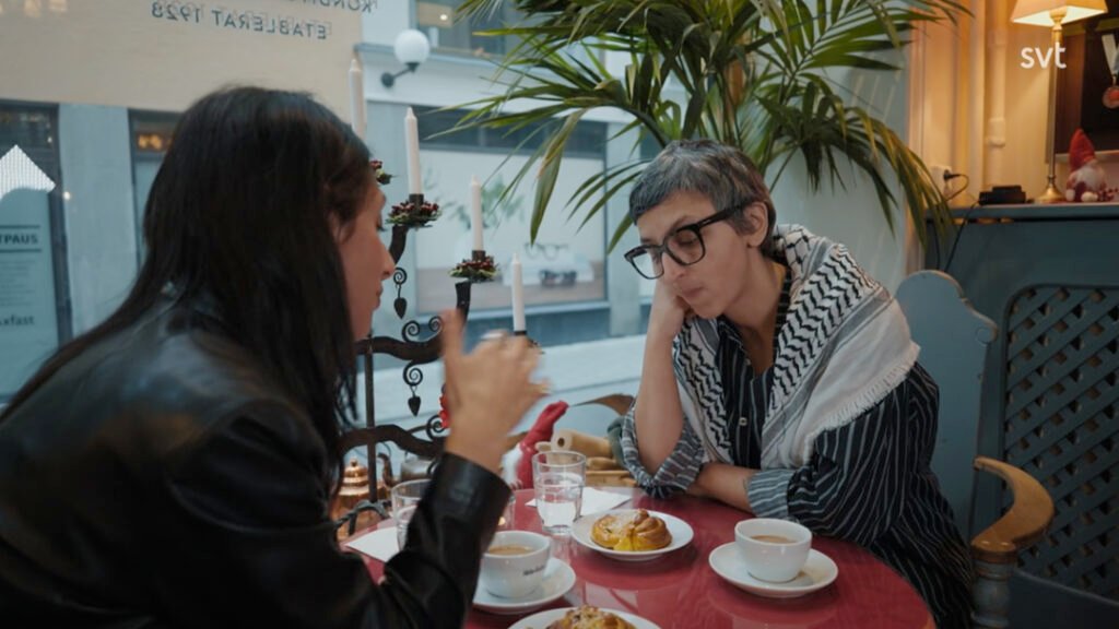Stiligt - Två personer sitter vid ett litet cafébord med kaffe och bakverk och samtalar vid ett fönster. Den ena vilar huvudet på handen och lyssnar.