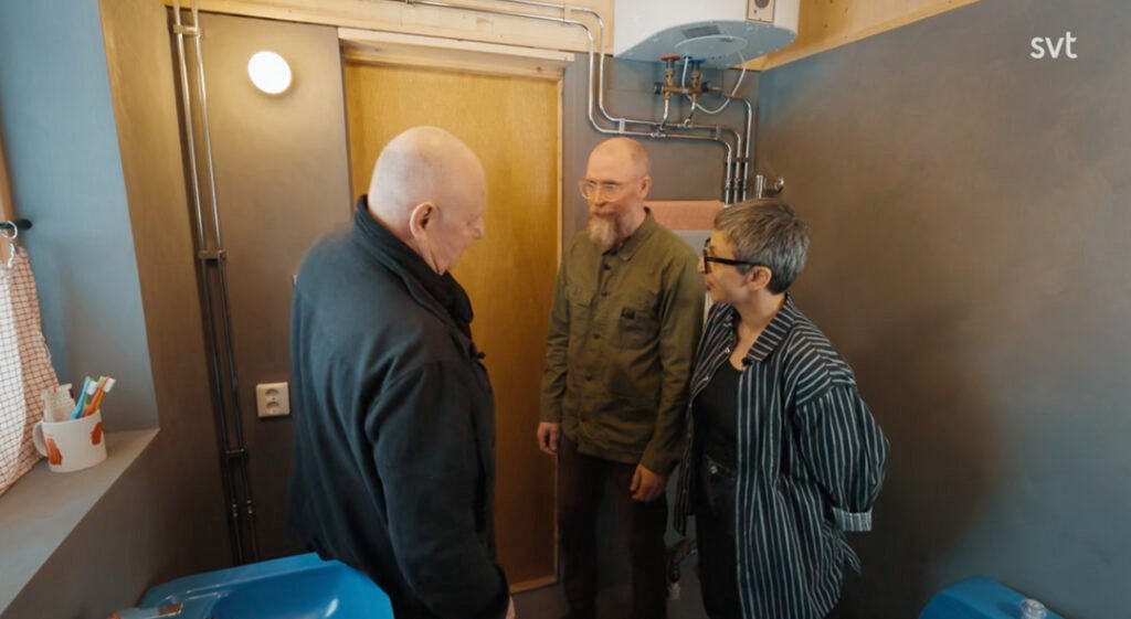 Stiligt - Tre personer står och pratar i ett litet, modernt badrum med synliga rör och en varmvattenberedare på väggen.