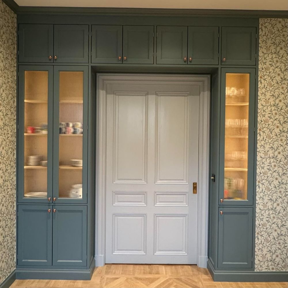Stiligt - Inbyggda gröna skåp med glasdörrar flankerar en central grå dubbeldörr och illustrerar den tidlösa charmen hos platsbyggda klassiska kök; på hyllorna står porslin uppställt, och tapeten runtom har ett mönster av lövrika rankor.