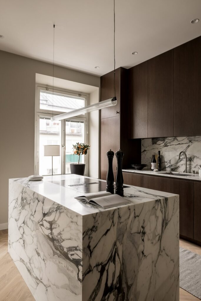 Stiligt - Ett modernt kök med skåp i mörkt trä, en stor köksö i marmor, en stänkskydd i marmor och minimalistisk inredning. Ett fönster släpper in naturligt ljus, och en växt står på bänkskivan bredvid diskbänken.