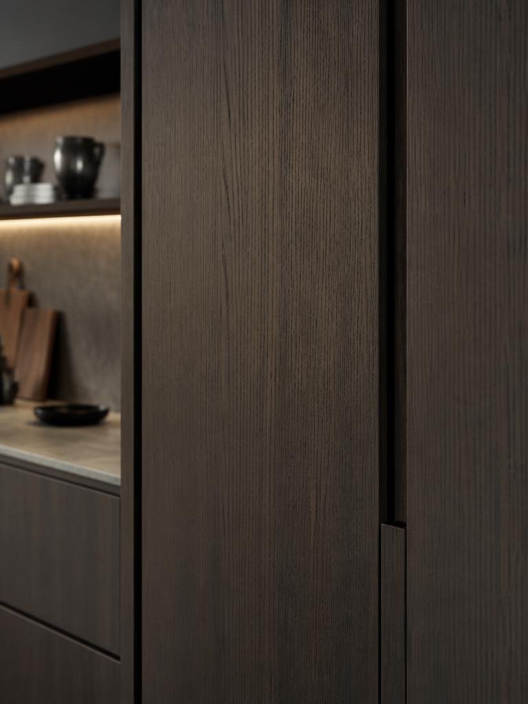 Stiligt - Närbild på ett modernt köksskåp i mörkt trä med en sömlös design. I bakgrunden syns hyllor med skålar och koppar, samt en arbetsbänk och skärbrädor.